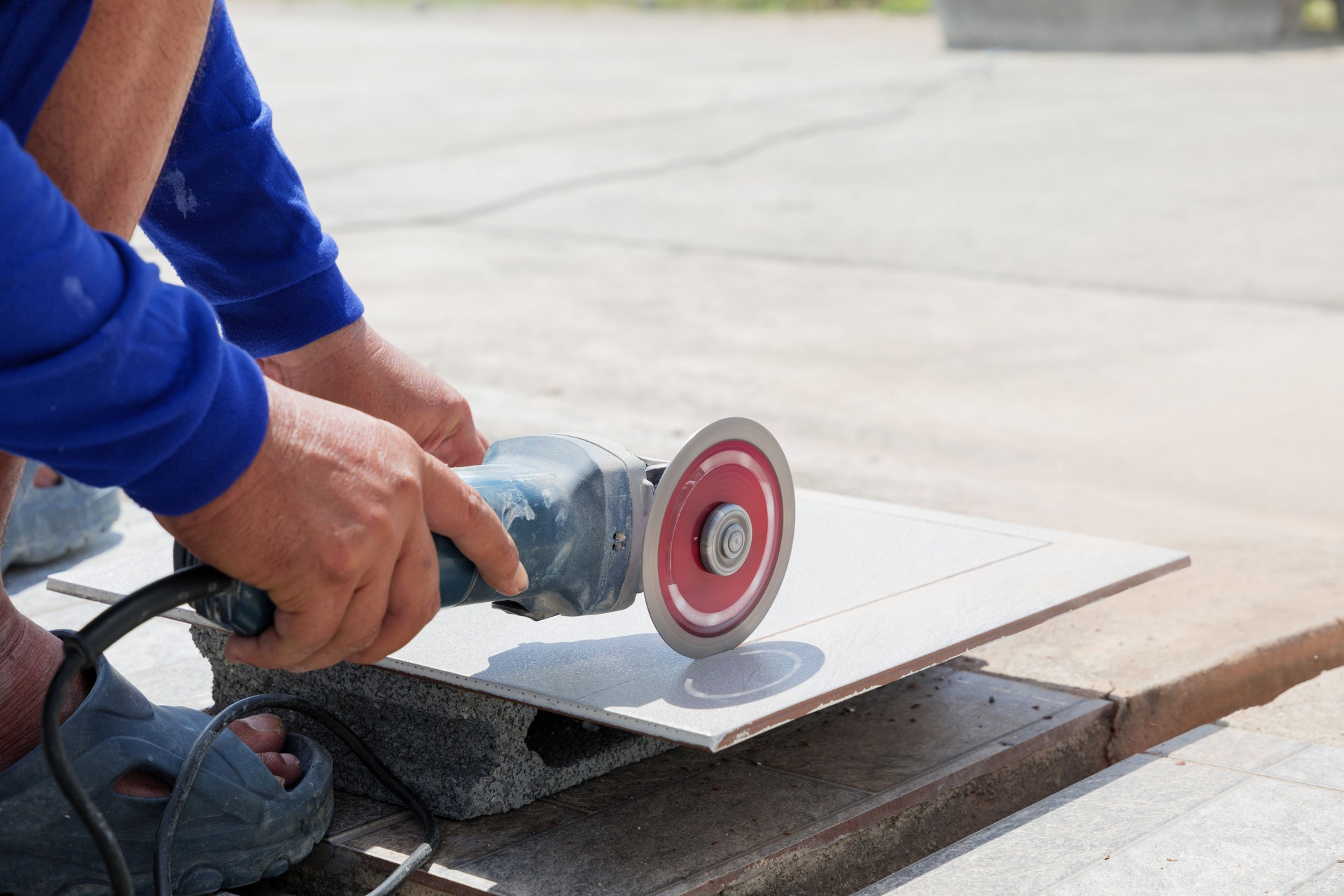 Worker in blue clothing uses an angle grinder to cut a gray tile on a piece of stone. Close-up of tool and process highlights precision and craftsmanship in tile cutting at construction site